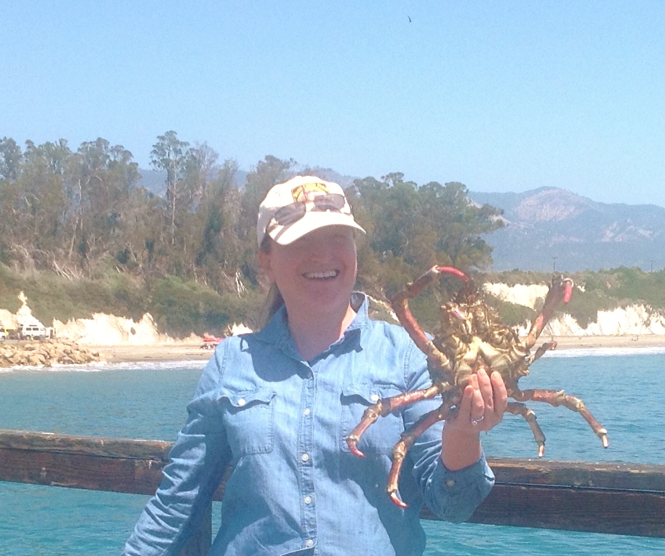 Dr. Quimby smiles as she holds up a large spider crab that a fisher (not her) caught on the Goleta Pier. Behind her is the shoreline and beach.