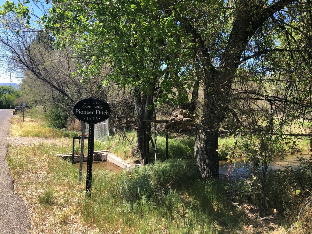 A sign reads "Historic Camp Verde Pioneer Ditch 1865" next to a small stream of water. 