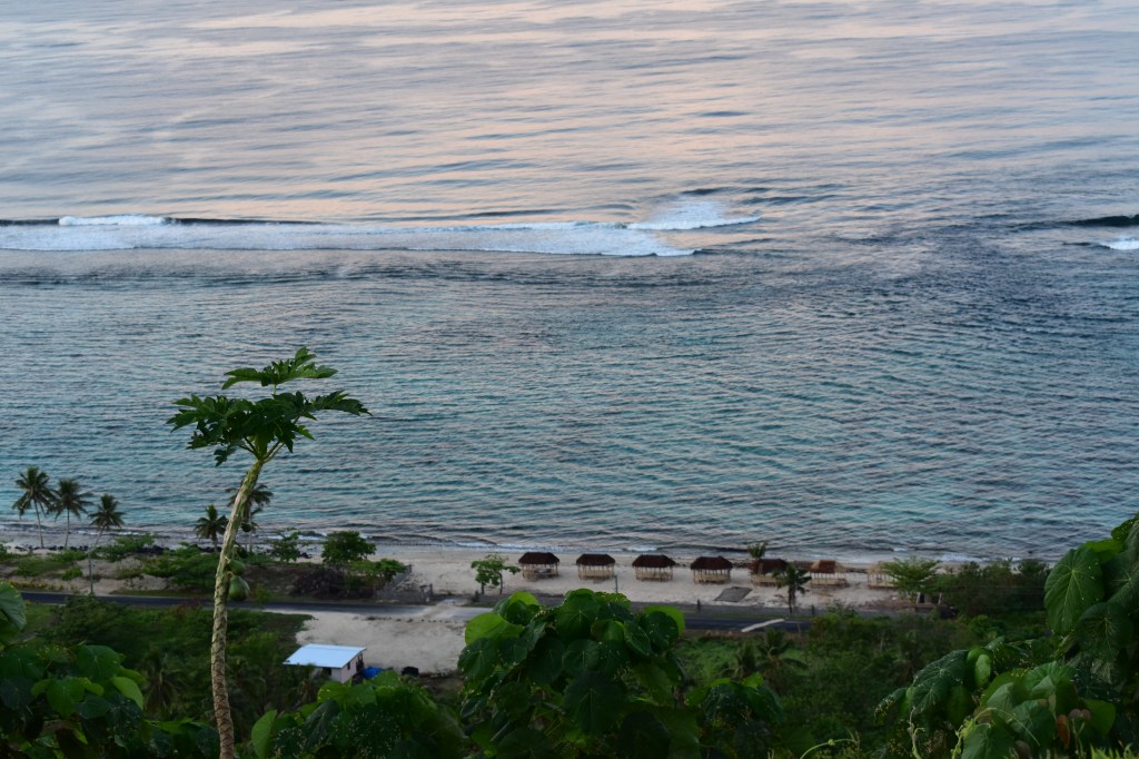 Lalomanu Beach, Upolu at sunset, looking down at beach fales from a high spot on a hill. The ocean water is pink and purple in the fading light.