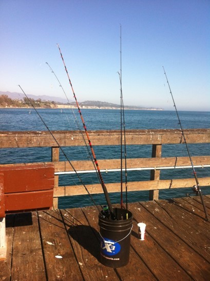 A bucket filled with fishing poles sits on the Goleta Pier with the ocean and shore in the background.